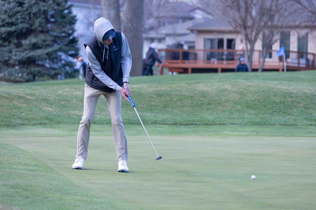A golfer in a hoodie and vest is poised to putt a golf ball on a green, with a blurred background of trees and spectators.