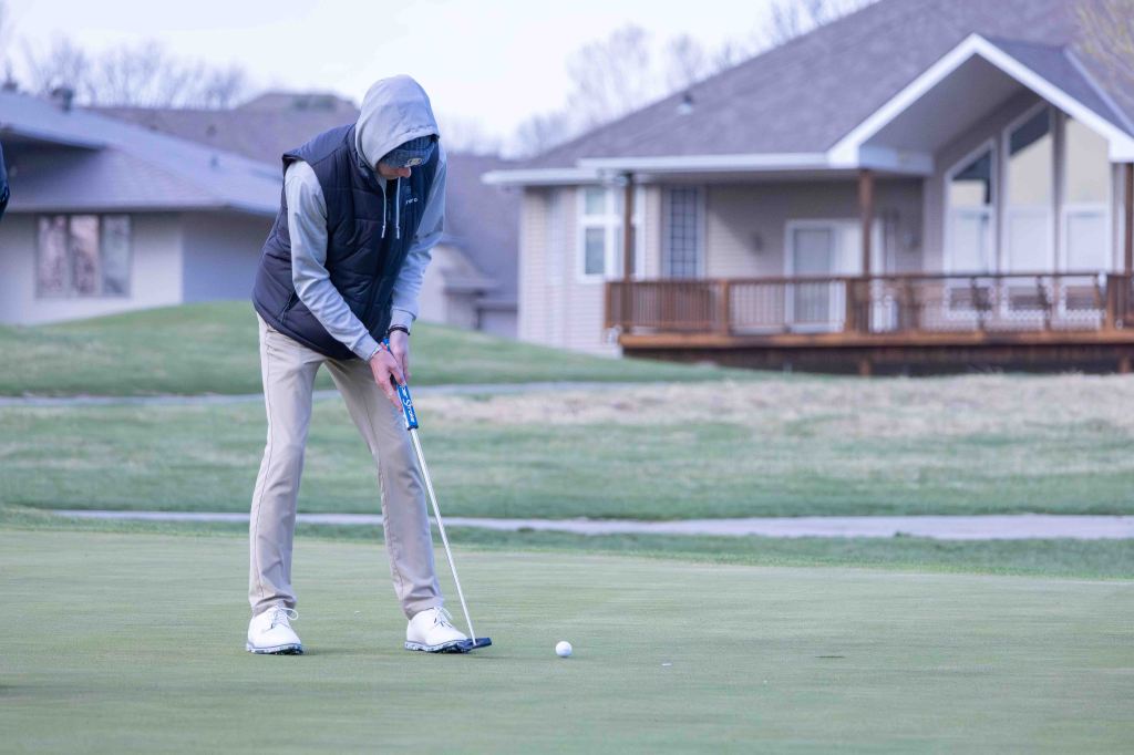 A golfer wearing a hooded jacket and khakis is putting on a green golf course with a house in the background.