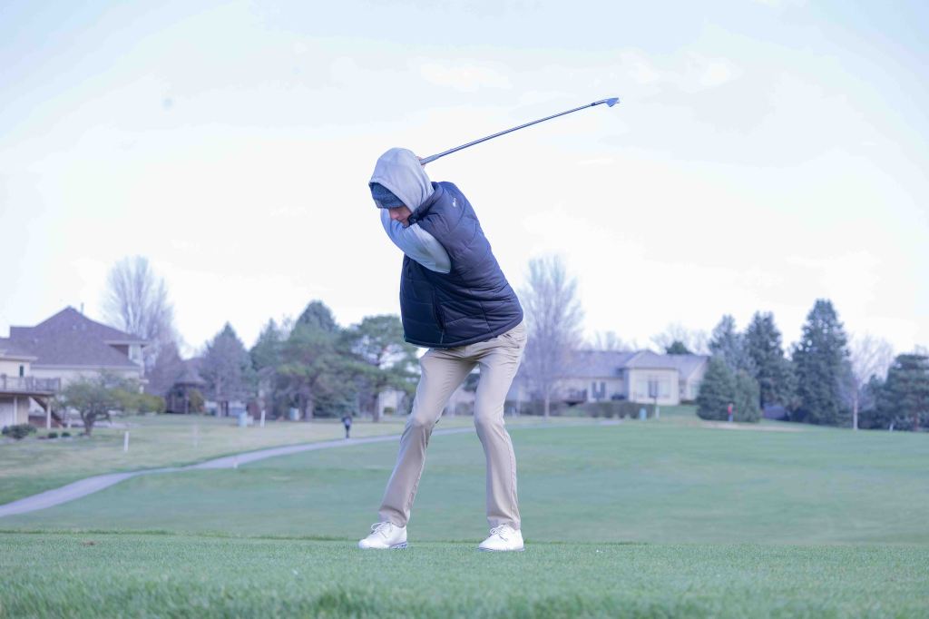 A golfer in a hoodie and vest is mid-swing on a golf course, with houses and trees in the background.