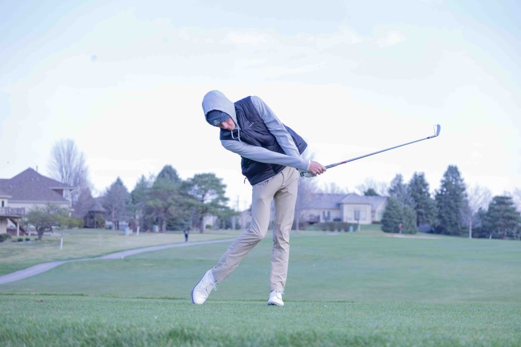 A golfer in a hoodie swings a golf club on a green golf course with houses and trees in the background.
