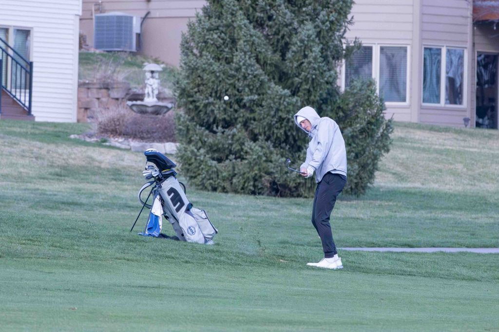 A golfer wearing a gray hoodie swings a club on the green grass of a golf course, with a golf bag in the background.