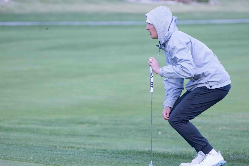 A golfer in a grey jacket and hoodie is crouched down on a green golf course, preparing to putt.