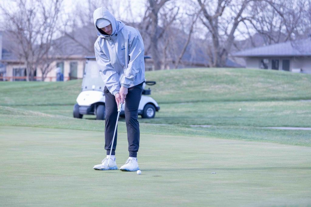 A golfer in a gray jacket and hood putting on a green golf course with a golf cart in the background.