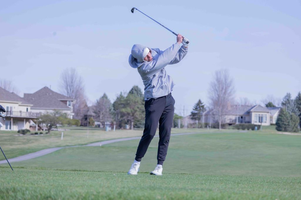 A golfer in a gray jacket and black pants swings a golf club on a grassy golf course with residential homes in the background.