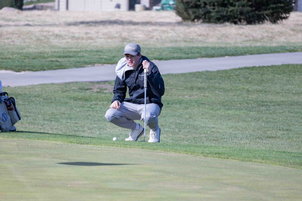 A golfer crouches on the green, analyzing a putt while holding a golf club, with a golf bag visible in the background.