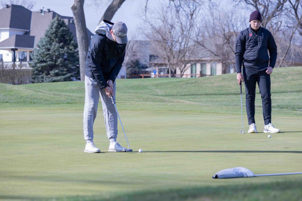 Two golfers on a putting green, one preparing to putt while the other observes.