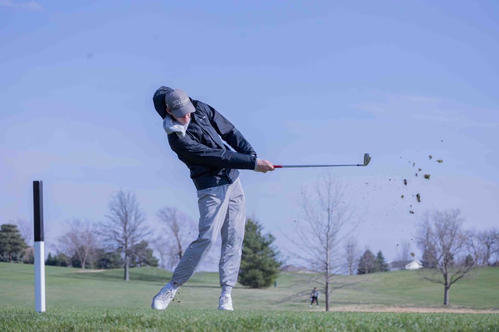 A golfer in a dark jacket and cap making a swing on a golf course, with grass flying up from the ground.