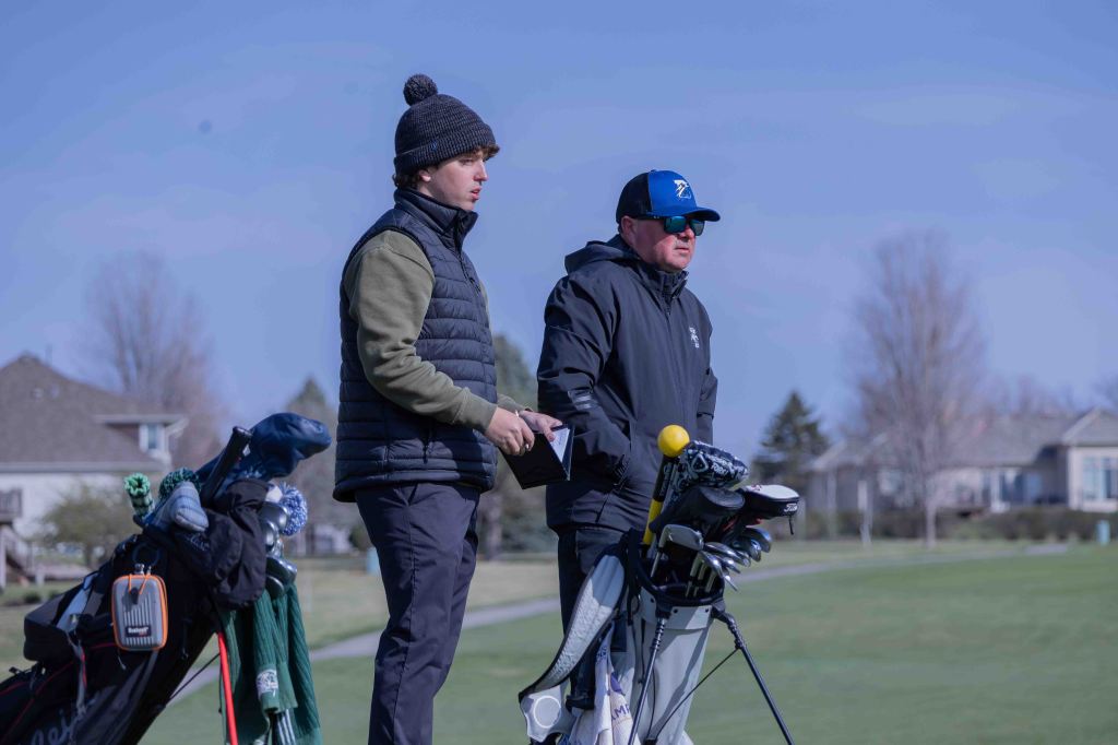 Two men standing on a golf course, one wearing a beanie and puffer vest, the other in sunglasses and a jacket, both beside golf bags.