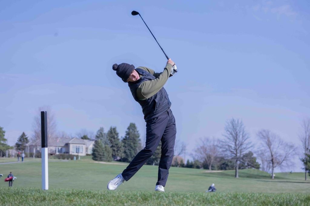 A golfer swinging a club on a green golf course, dressed in a black beanie, a dark vest, and pants, with trees and a house in the background under a clear blue sky.
