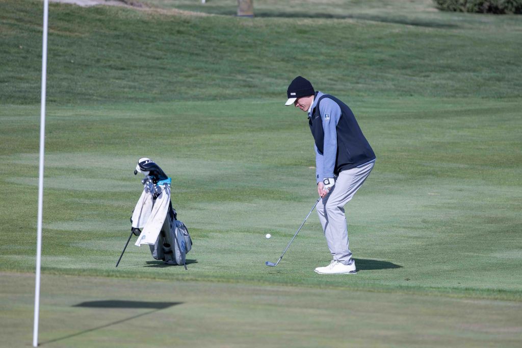 A golfer in a beanie and vest preparing to take a shot on the green, with golf clubs in the background.