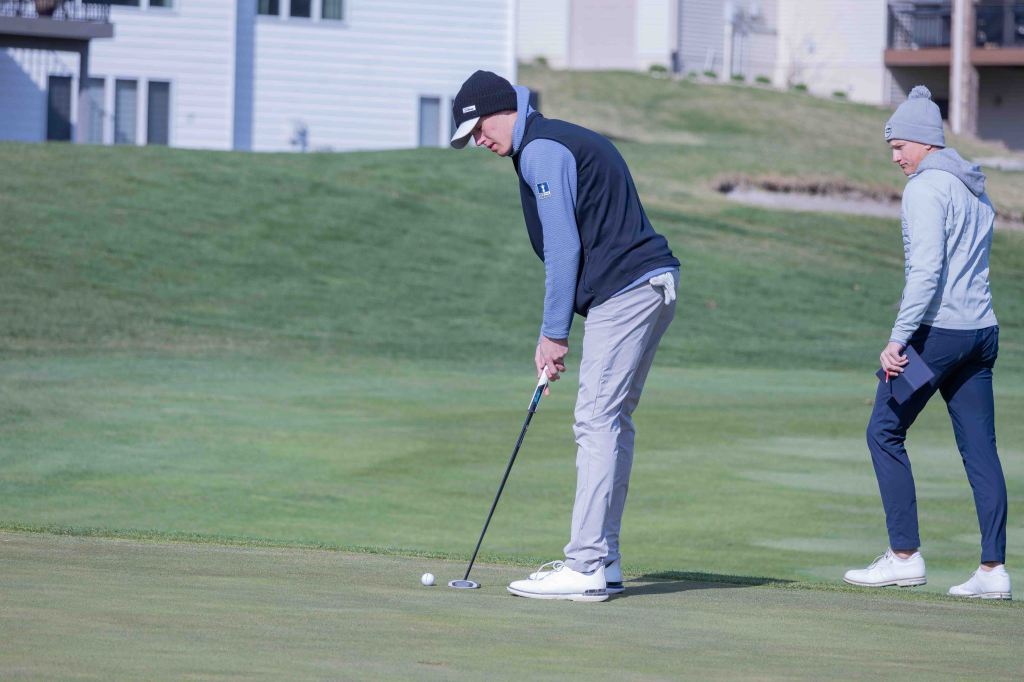 A golfer preparing to putt on a green, with another person observing in the background, dressed in casual golf attire.