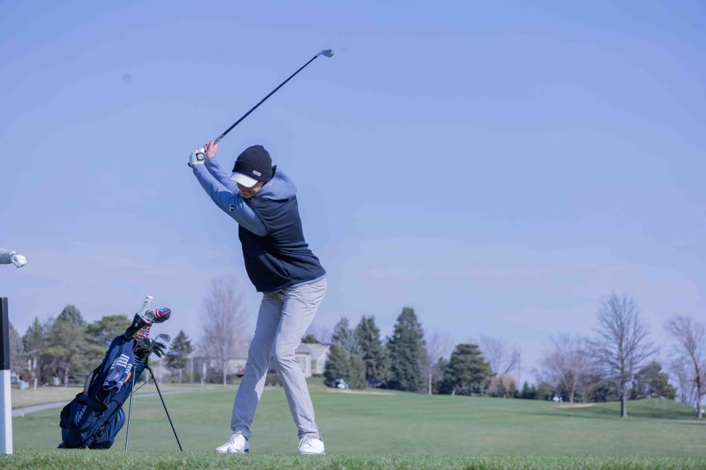 A golfer in a black and gray outfit is taking a swing on a green golf course with trees in the background.