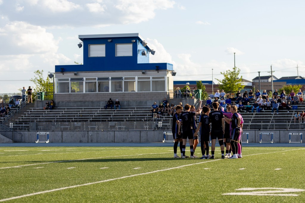 A soccer team in black uniforms huddles together on the field, with a blue press box and spectators in the background.