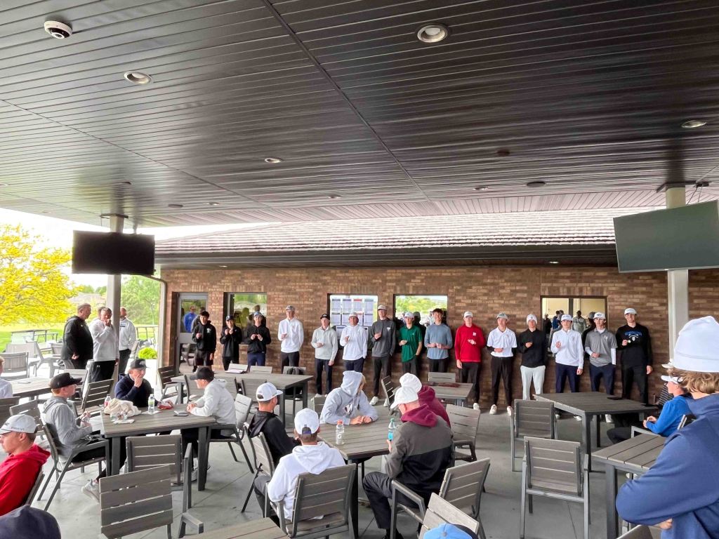 A group of young athletes stands in a semi-circle, engaging in a presentation or gathering at an outdoor patio area with tables and chairs.