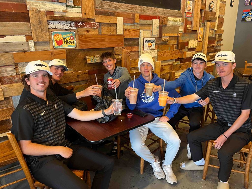 Group of six young men in a casual setting, sitting around a table, holding drinks and smiling, with wooden wall decor in the background.