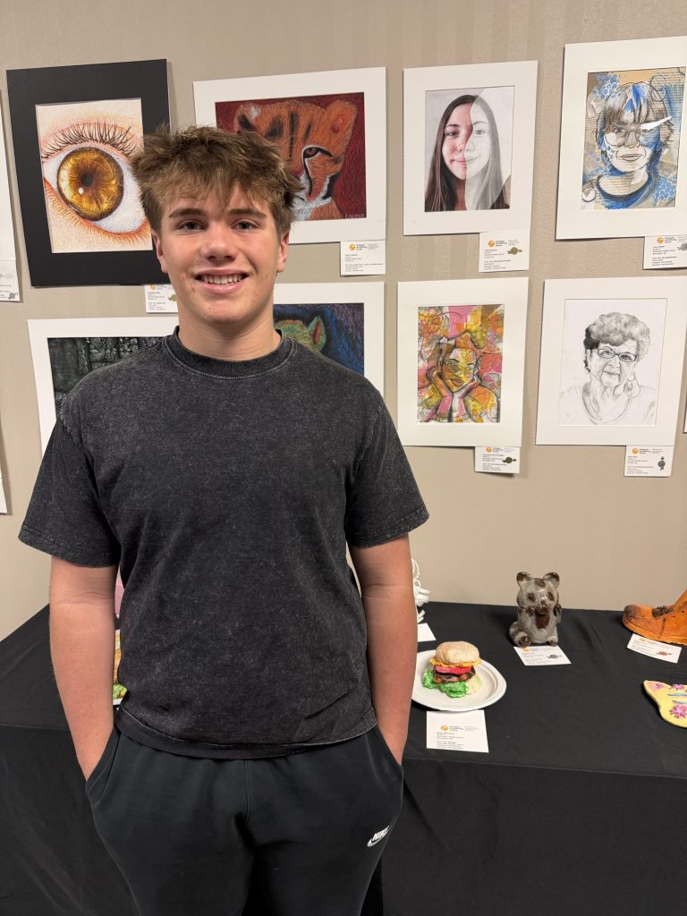 A young man with short brown hair is standing in front of various artworks displayed on a wall. The art includes portraits and animal illustrations. A table in front features various artistic creations, including a food sculpture.