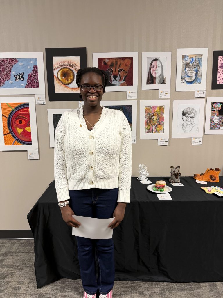 A young woman stands smiling in front of a display of artwork at an exhibition. She is wearing a white sweater, dark jeans, and holding a piece of paper. Behind her, colorful framed artworks are hanging on the wall, and a table with various art pieces is visible to her right.