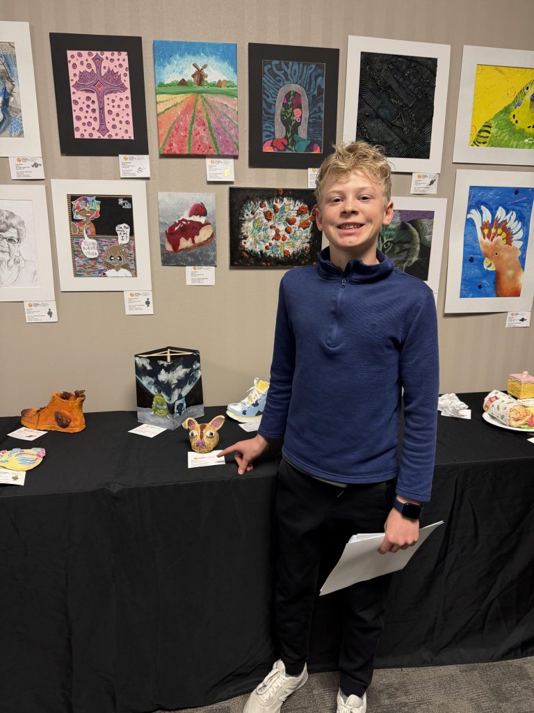A smiling boy stands in front of a display of student artwork at an exhibition. Various colorful paintings are hung on the wall behind him, and unique art pieces are displayed on a table in front.