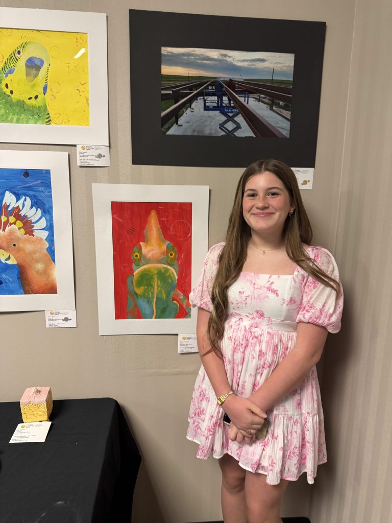 A young girl with long brown hair smiles while standing next to a display of colorful artwork on the wall behind her, including paintings of a parrot, a fish, and an abstract scene.