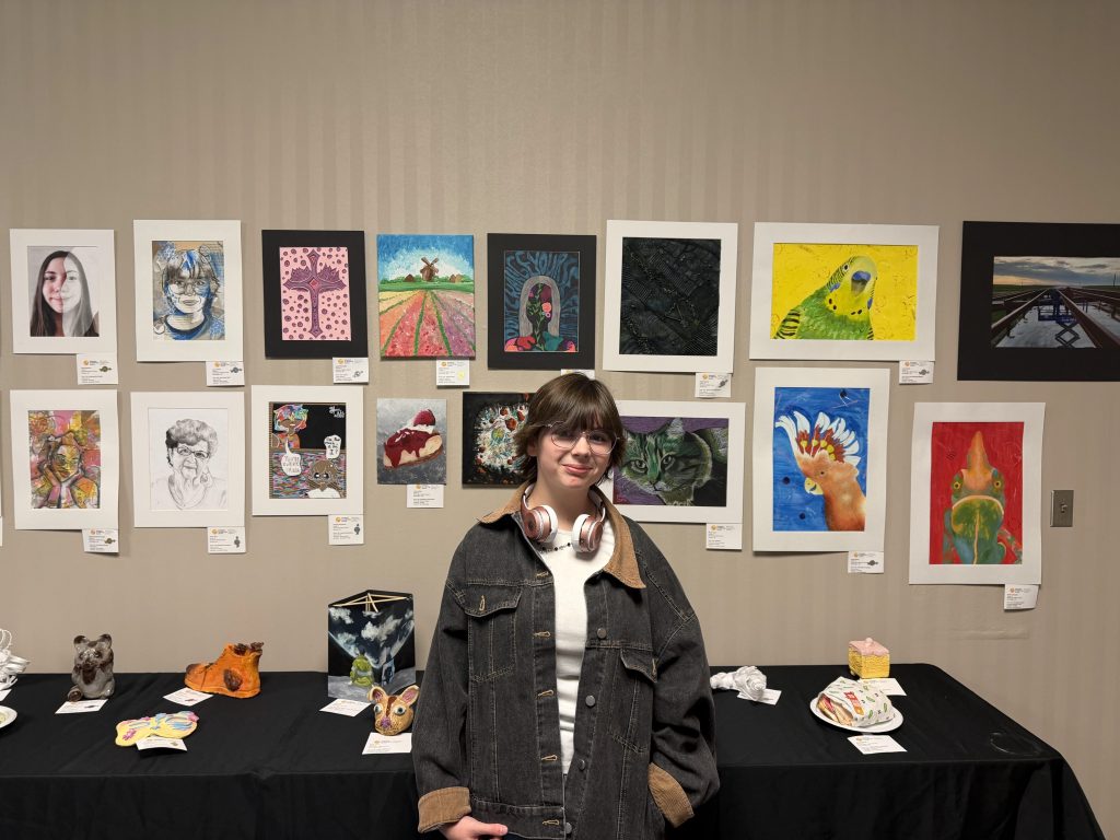 A young person stands in front of a wall displaying various pieces of colorful artwork, including portraits, animals, and abstract designs. There is a black table in the foreground with art pieces and decorations.