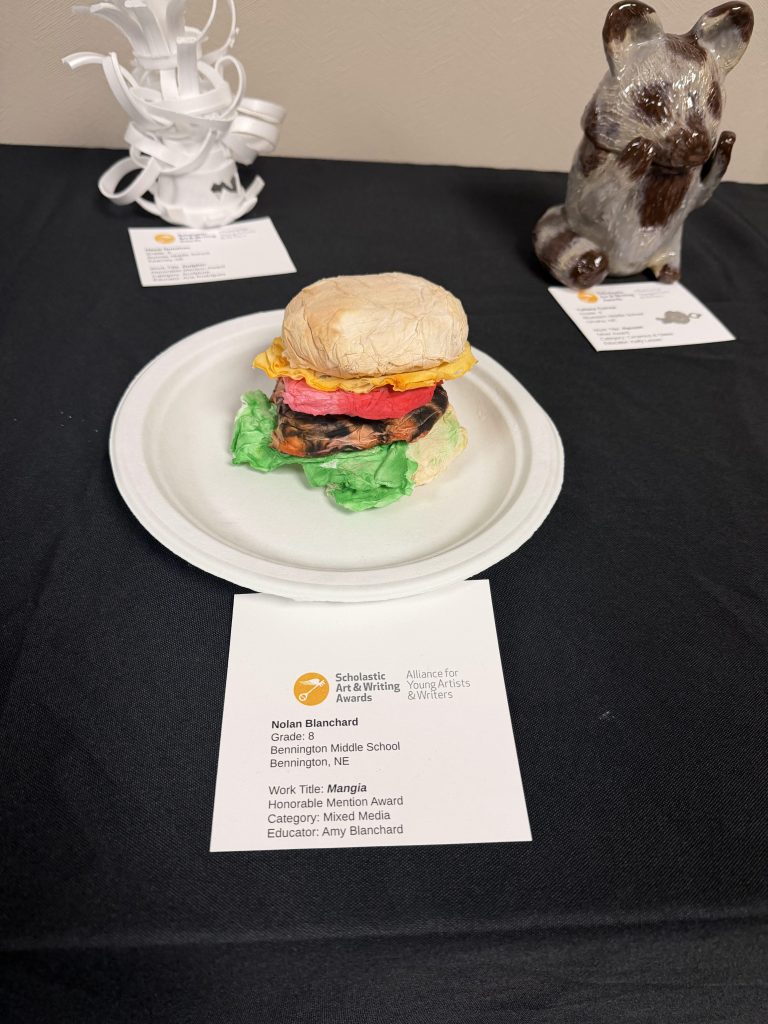 A mixed media sculpture of a hamburger made from colorful materials on a paper plate, displayed at an art show. The artwork is titled 'Mangia' by Nolan Blanchard, an eighth grader from Bennington Middle School, Nebraska.