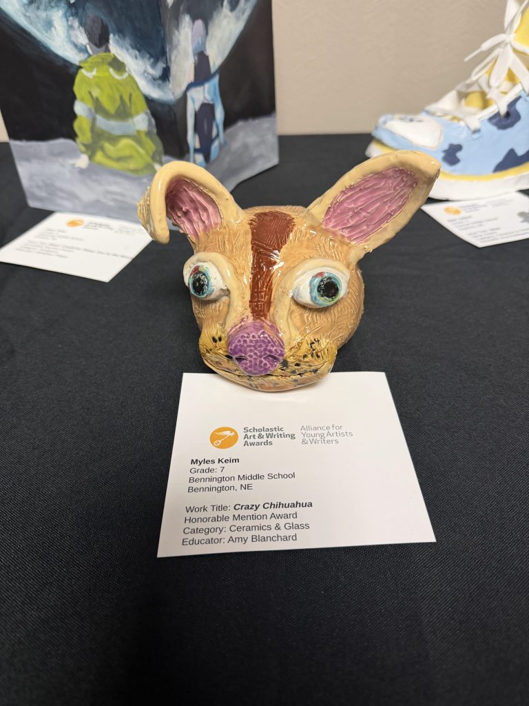 A ceramic sculpture of a whimsical chihuahua head with large ears and colorful features, displayed on a black table along with an awards card for the artist, Myles Keim, from Bennington Middle School.