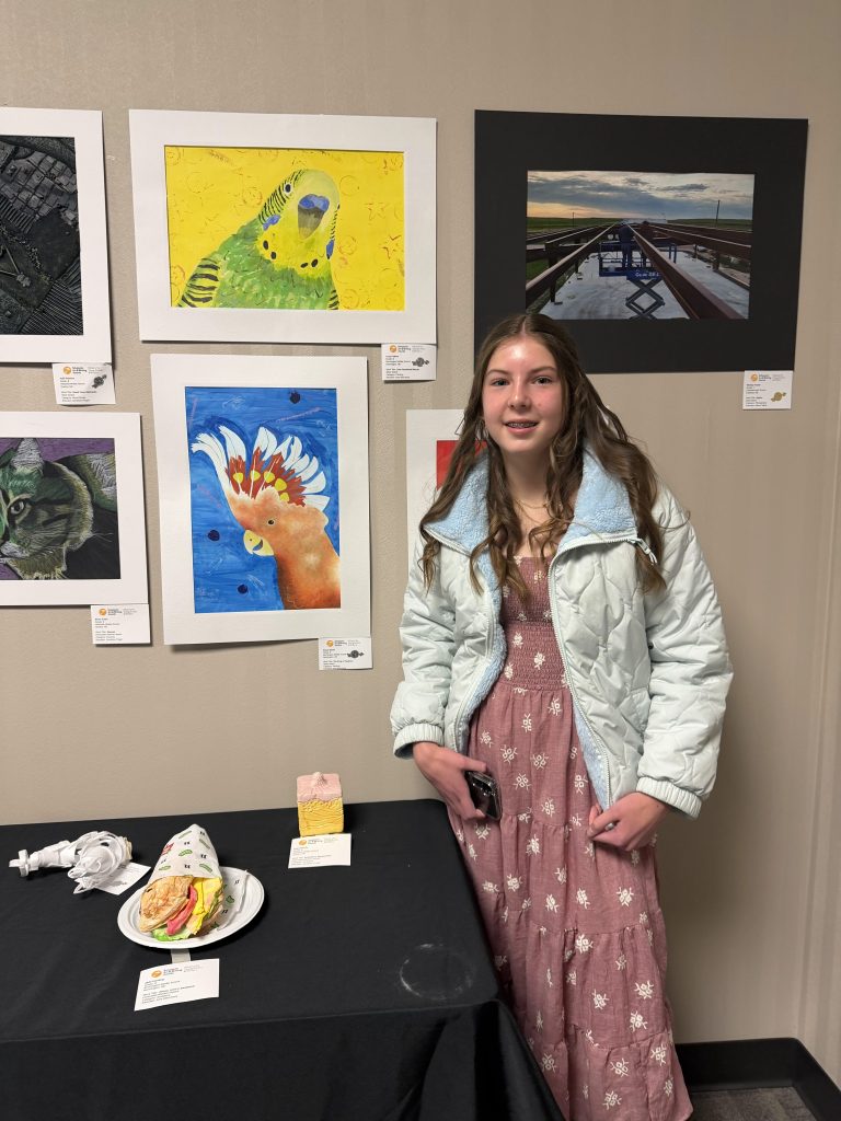 A young girl stands beside an art display featuring colorful paintings, including a parakeet, a rooster, and a cat. She wears a light blue jacket and a pink dress, holding a phone, with a table in front showing a faux sandwich and a cake.
