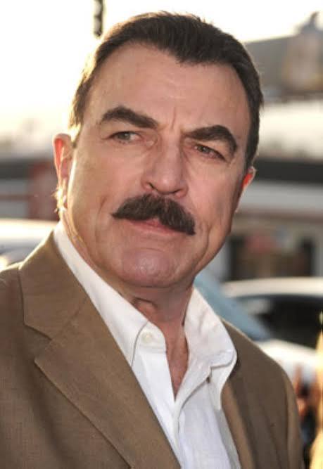A close-up portrait of a man with a thick moustache, wearing a beige suit jacket and a white shirt, looking serious against a blurred outdoor background.
