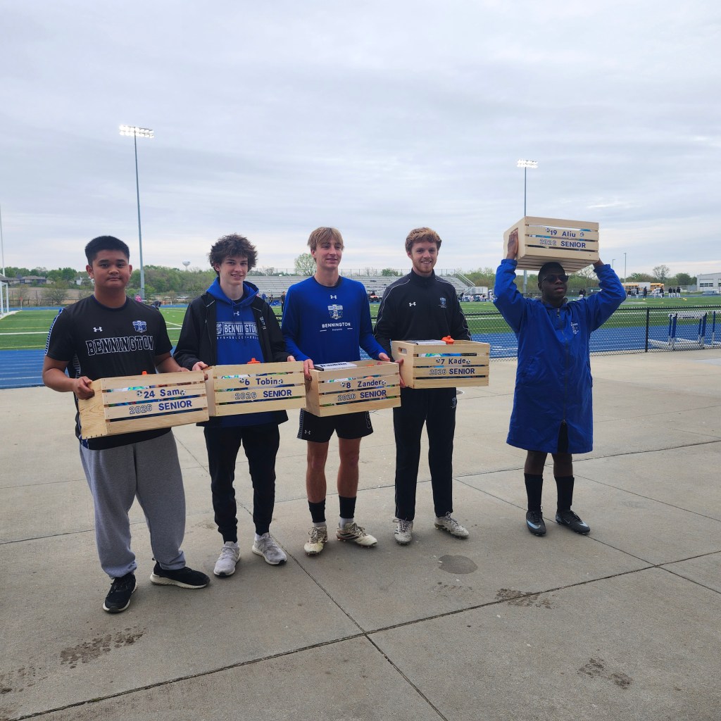 Five male high school seniors pose outdoors, holding wooden boxes with their names and 'SENIOR' written on them, in front of a sports field.