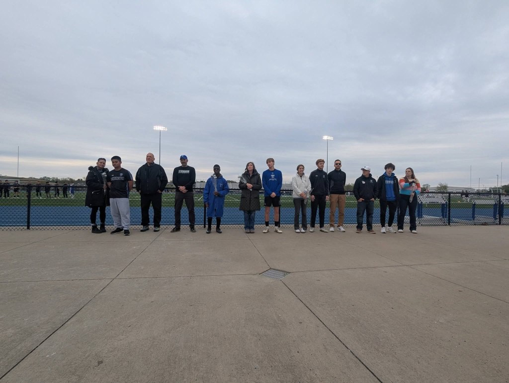 A group of people standing in a line on a track field, facing forward. They are dressed in various athletic and casual clothing, with a cloudy sky in the background.