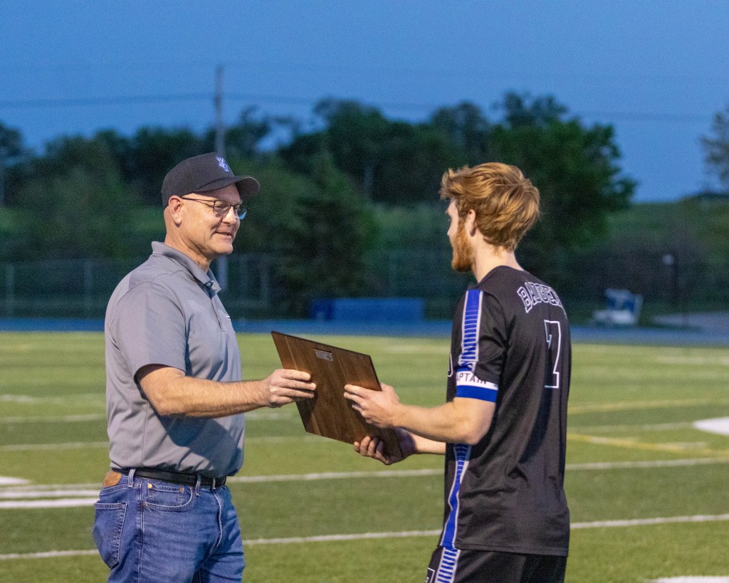 A man handing an award plaque to a young athlete on a soccer field during an evening ceremony.