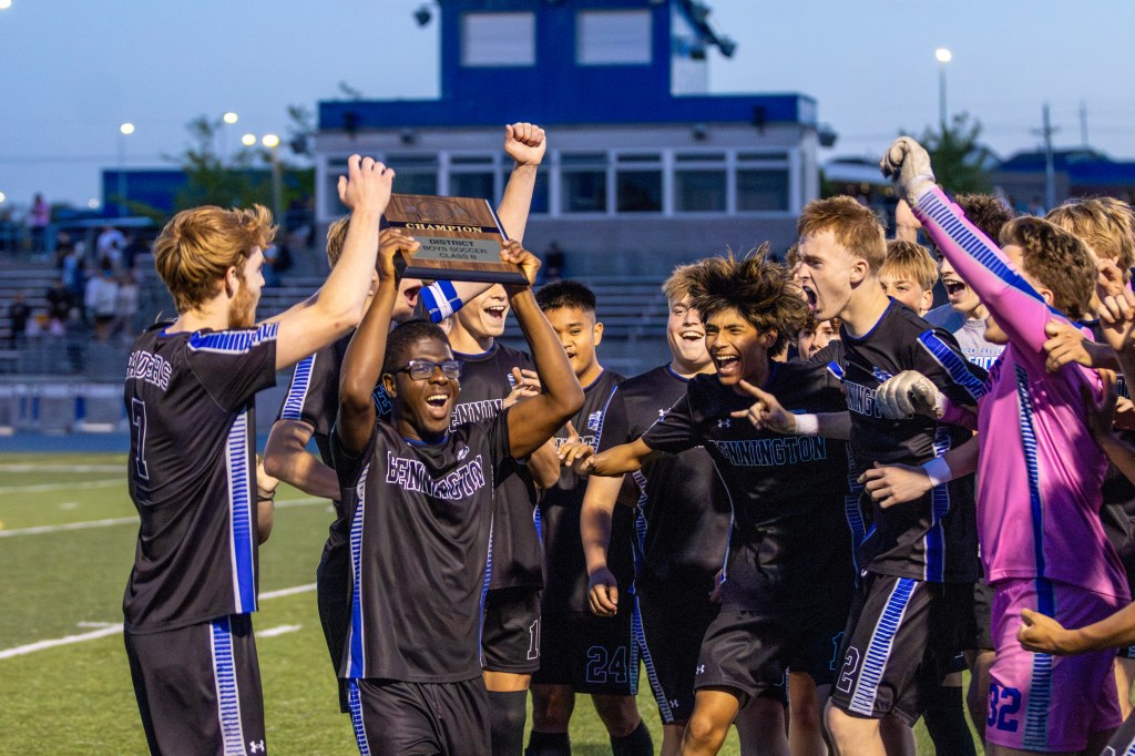 A group of excited soccer players celebrating and holding a championship plaque on the field at dusk.
