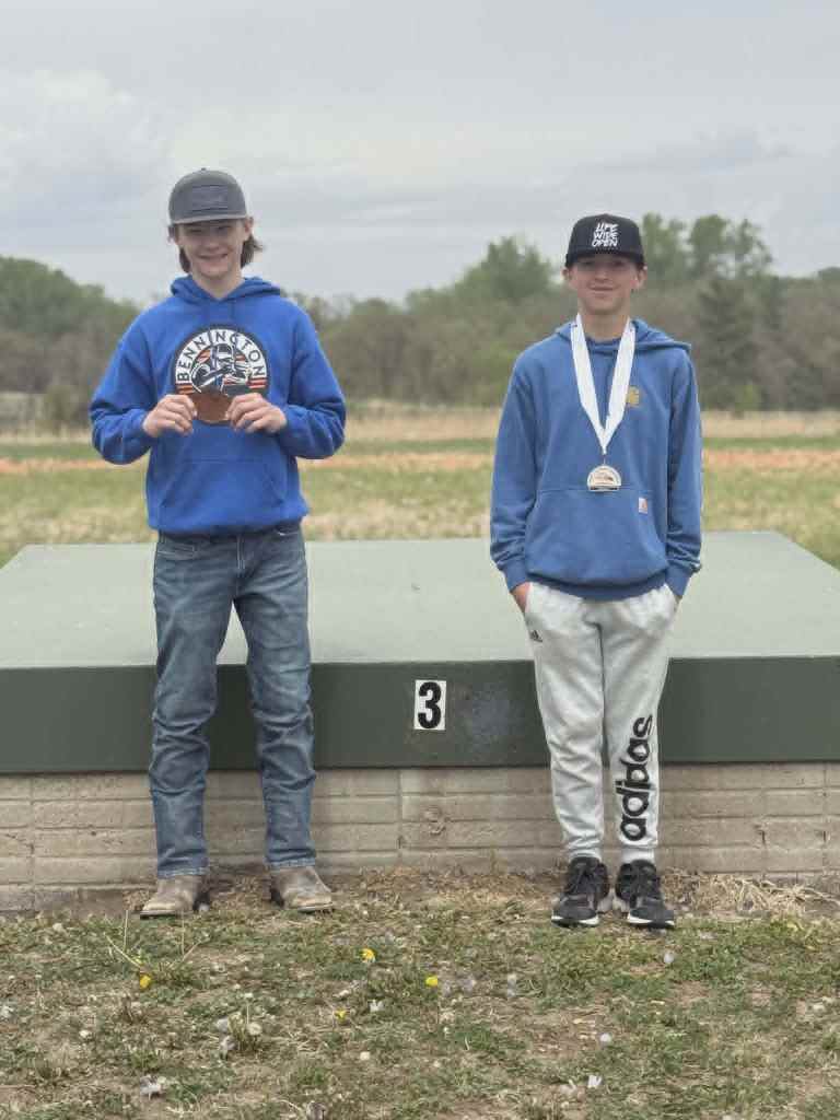 Two boys posing on a podium after a competition, one holding a medal, with a grassy background and cloudy sky.