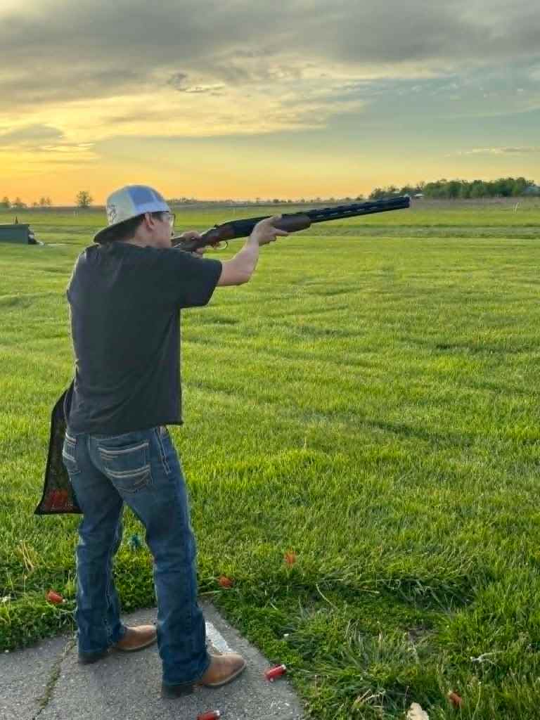 A person aiming a shotgun while standing on a grassy field during sunset.