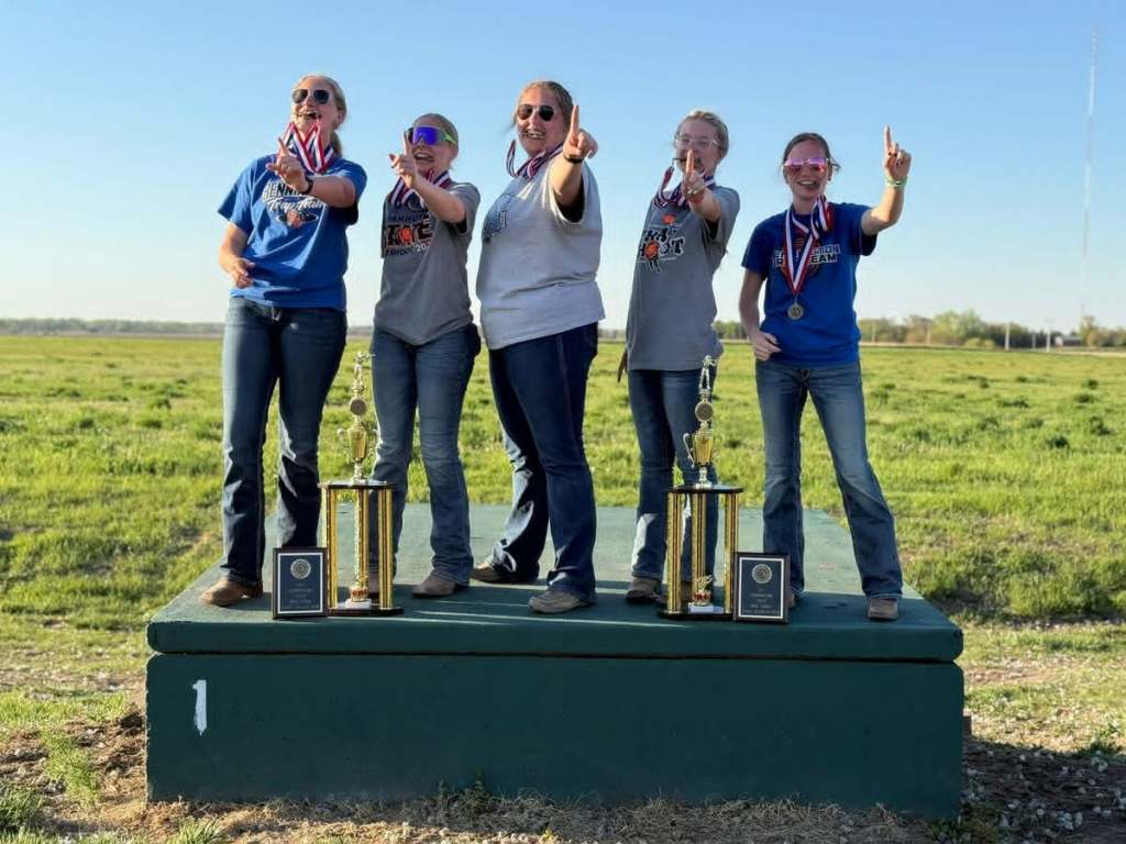 Five female winners celebrating on a podium, holding trophies and medals, outdoors in a grassy field, with each person showing a 'number one' gesture.