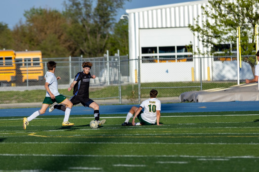 A soccer match in progress with players in action on the field. One player in black is kicking the ball while two players in white and green are positioned nearby, one sitting on the ground.