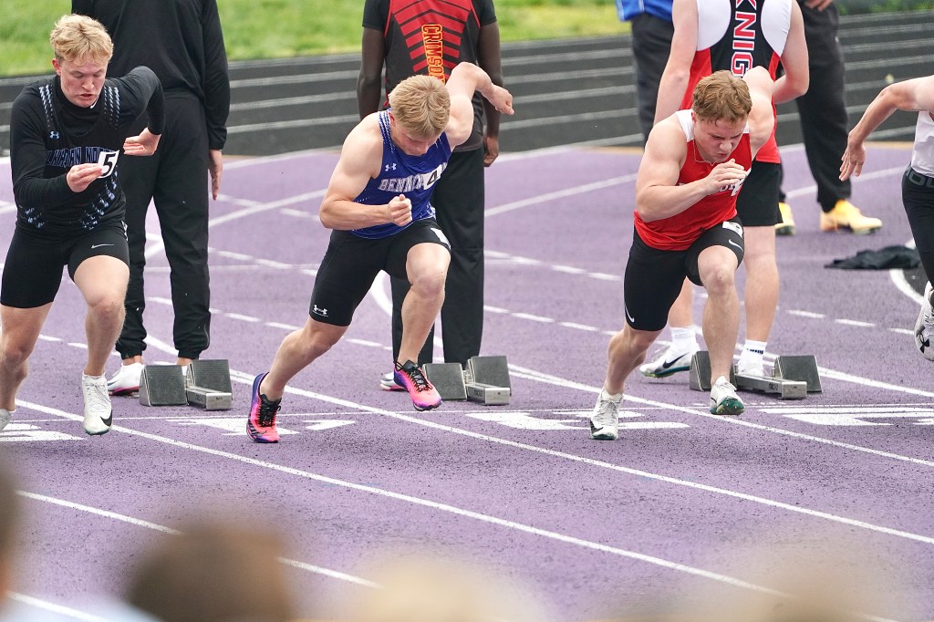 A group of male sprinters in athletic uniforms starting a race on a purple track, with some runners in motion and a spectator blurred in the foreground.