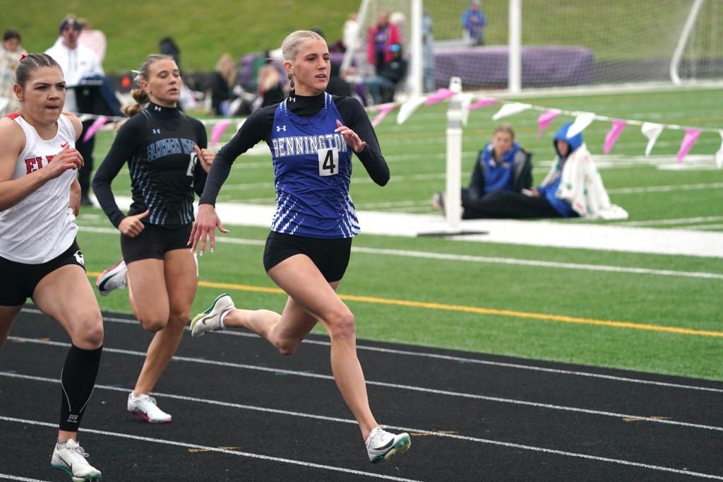 A group of female athletes competing in a sprint race on a track field, with one runner in a blue uniform leading the way.