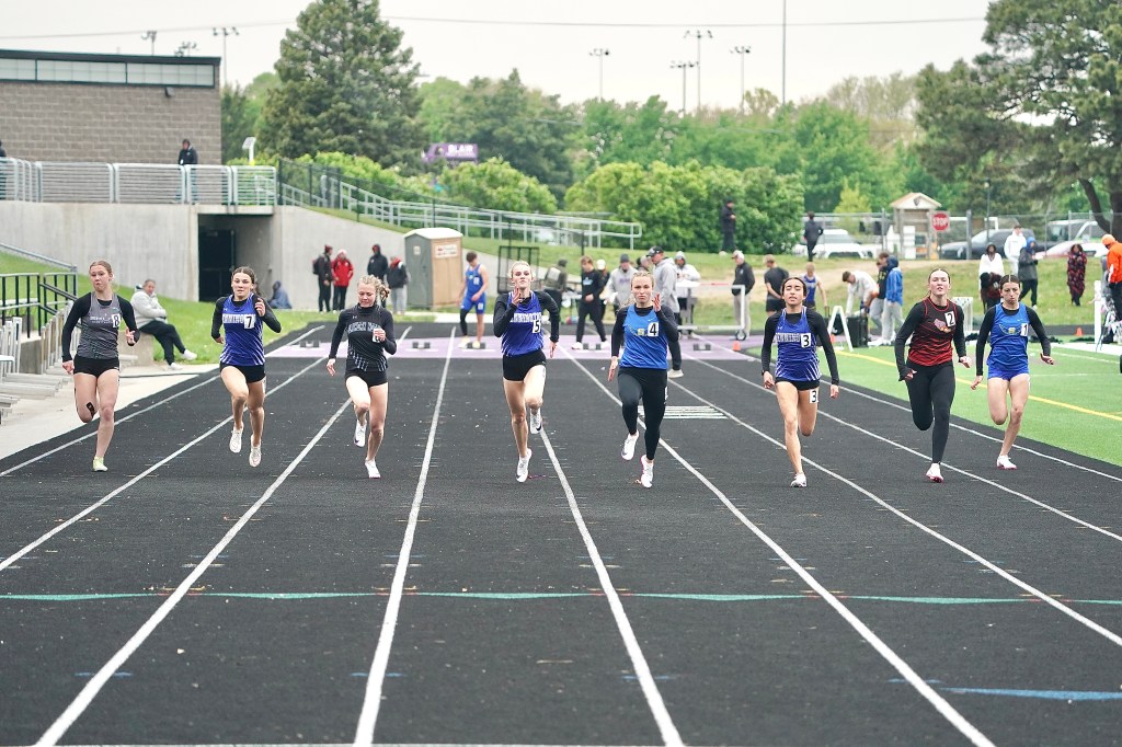 A group of female athletes sprinting down a track during a competitive race on an outdoor field.