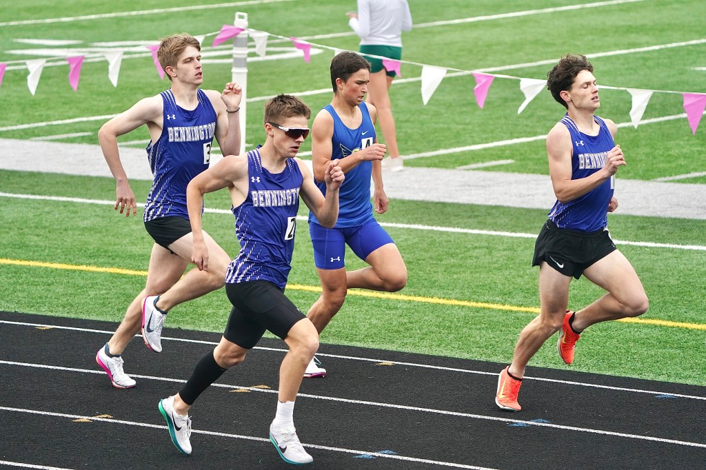 Four male track athletes running in a race on a field, wearing athletic uniforms in blue and black.