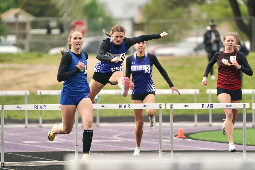 A group of female track athletes competing in a hurdle race, with one athlete leaping over a hurdle in the foreground while others are running behind her.