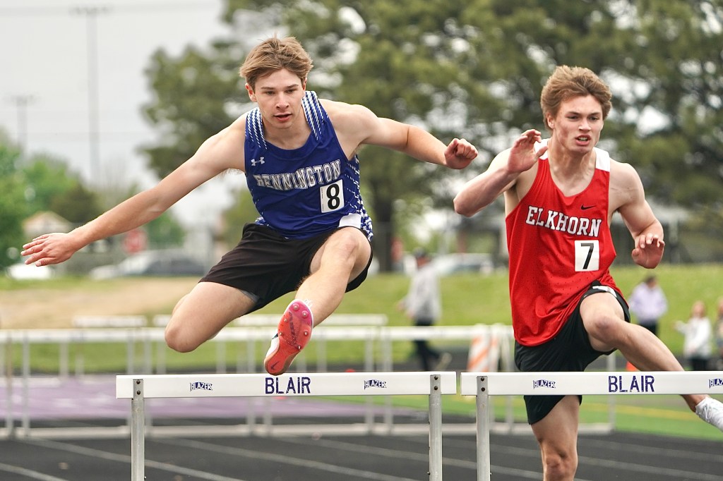 Two male track athletes competing in a hurdles race, one wearing a blue jersey with 'Bennington' and the other in a red jersey with 'Elkhorn'.