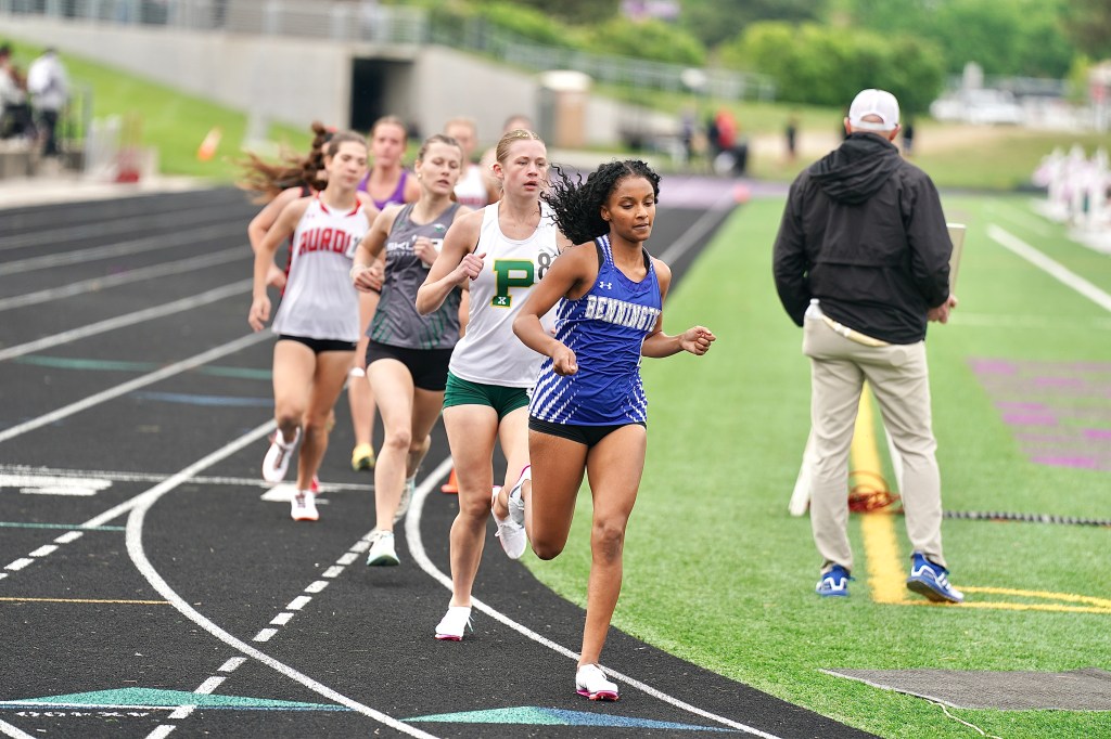 A group of female runners competing on a track, with one athlete in a blue uniform leading the race. The scene conveys a dynamic athletic competition with a grassy area in the background.