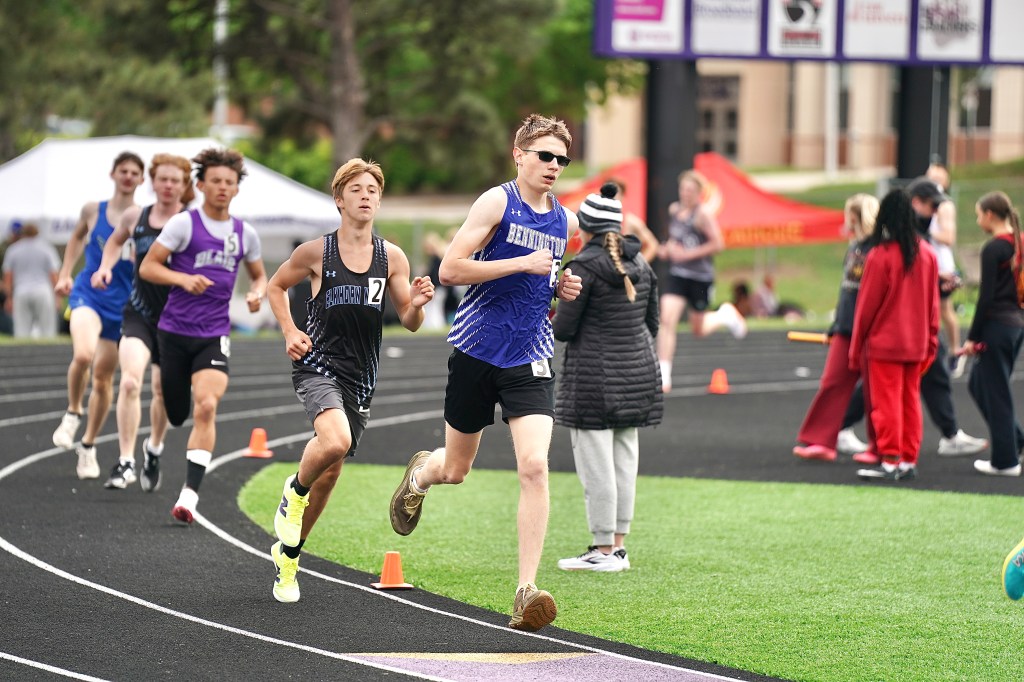 A group of high school track runners competing on an outdoor track, with a focus on two athletes in the foreground as they sprint during a race.