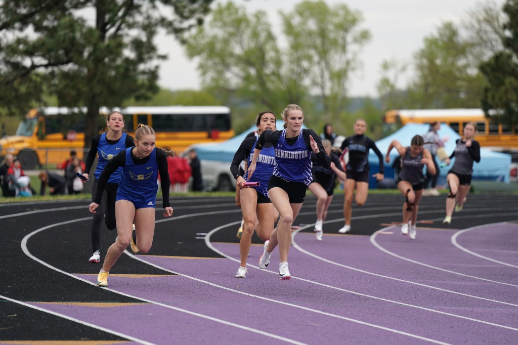 A group of female track athletes sprinting on a purple and black running track during a race. Trees and a school bus are visible in the background.