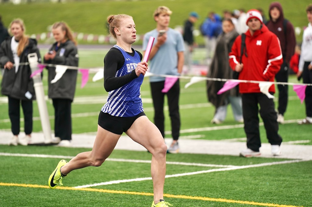 A female athlete running on a track while holding a relay baton, with spectators in the background.