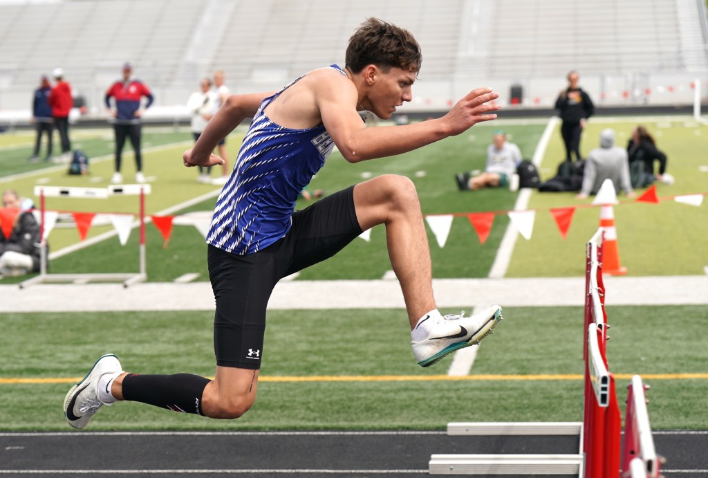 A male athlete competing in a hurdle race, mid-jump over a hurdle, wearing a blue and white athletic uniform on a track field.