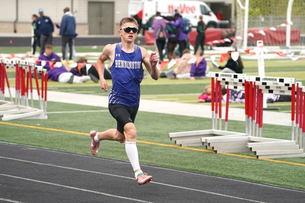 A male athlete in a blue tank top with 'BENNINGTON' printed on it, wearing sunglasses, runs on a track with hurdles in the foreground. Other athletes and spectators can be seen in the background.