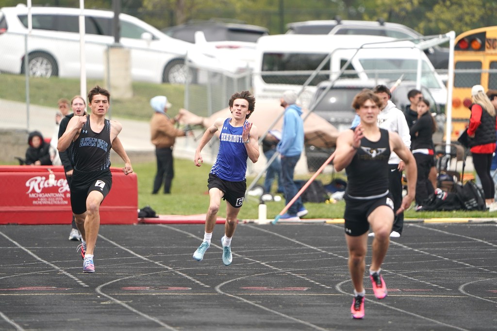 Three high school runners competing in a sprint race on a track, with spectators and vehicles in the background.