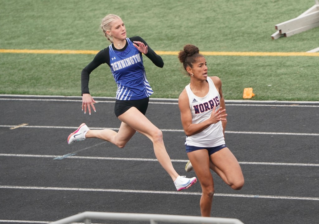 Two female track athletes competing in a race on a running track, one in a blue jersey and the other in a white jersey.